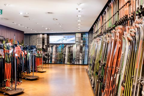 Ein Ladengang mit Regalen voller bunter Skier und Stöcke auf beiden Seiten, vor einem Holzboden. Am anderen Ende hängt unter Deckenleuchten ein großes Bild von schneebedeckten Bergen.