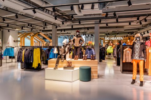 Das Bild zeigt das Innere eines Skibekleidungsgeschäfts mit Schaufensterpuppen in Winterkleidung und -ausrüstung. Das Geschäft stellt bei hellem Licht verschiedene Jacken, Hosen und Accessoires aus. Auf einem Schild steht „SKI HERREN“.
