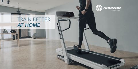 Persona corriendo en una cinta de correr Horizon Fitness en una habitación moderna y espaciosa con pisos de madera y grandes ventanales. Junto al logotipo de Horizon Fitness está el texto "Entrena mejor en casa".