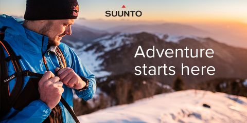 Una persona con una chaqueta azul y un gorro negro revisa su dispositivo de muñeca mientras está de pie en una montaña nevada al atardecer. El cielo es naranja y hay montañas al fondo. El texto dice: "La aventura comienza aquí".