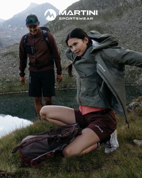 Dos personas con equipo de exterior y mochilas están de pie junto a un lago de montaña. Una está arrodillada ajustándose la chaqueta, la otra está de pie sonriendo. Al fondo, terreno rocoso y cielo nublado. En la parte superior de la imagen se ve "MARTINI SPORTSWEAR".