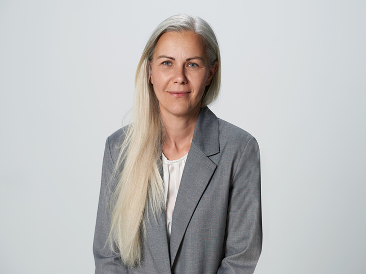 Una persona con cabello largo, liso y claro lleva un blazer gris y una blusa blanca. Está sentada mirando a la cámara con una expresión facial neutra frente a un fondo claro y sencillo.