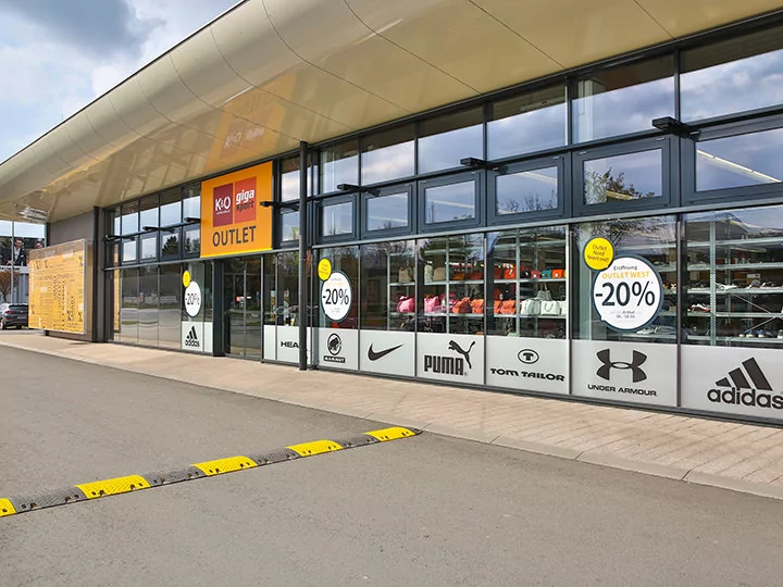 Vista exterior de una tienda outlet de deportes con grandes ventanales que muestran varios logotipos de marcas como Puma y Adidas. Los carteles en las ventanas anuncian un 20% de descuento en muebles, cortinas y textiles para el hogar. El edificio destaca por su diseño moderno.