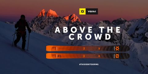 Un esquiador se encuentra en la cima de una montaña nevada al atardecer, recortado contra el cielo como una silueta. En el fondo se ven las cumbres iluminadas. En primer plano se ven esquíes Völkl de color naranja. El texto dice "Por encima de la multitud" con los hashtags #THISISSKTOURING.