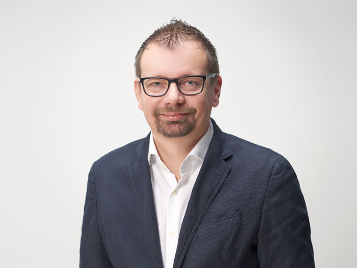 Un hombre con pelo corto y gafas lleva un blazer azul marino sobre una camisa blanca. Sonríe y está de pie frente a un fondo claro y sencillo.