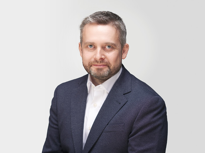Un hombre con pelo corto gris y barba lleva una chaqueta oscura y una camisa blanca. Está sentado frente a un fondo blanco sencillo y mira directamente a la cámara con una expresión facial neutra.