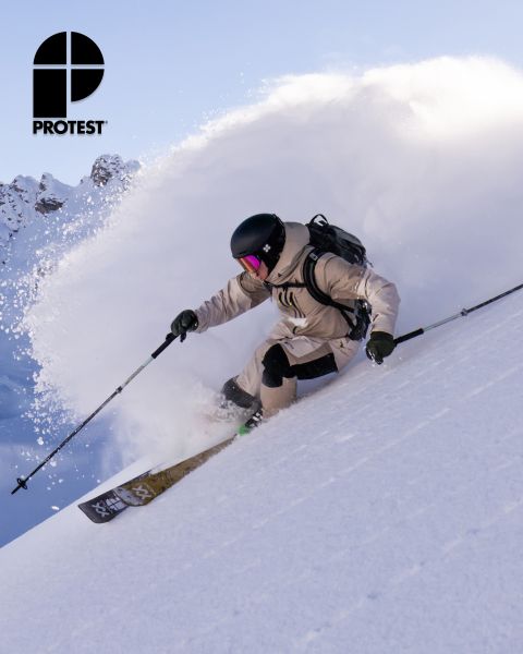 Un esquiador con equipo y casco beige desciende por la nieve polvo en una ladera empinada, dejando una nube de nieve. El logo "PROTEST" se ve en la esquina superior izquierda. En el fondo, se ven un cielo despejado y montañas nevadas.
