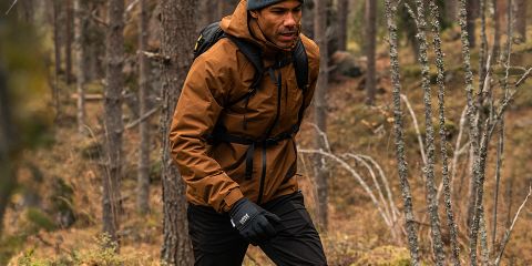 Hombre con chaqueta marrón, pantalones negros y gorro gris camina por un denso bosque. Lleva guantes negros y una mochila. El bosque tiene árboles altos y hojas caídas. Logotipo de "Rukka" en la parte superior.