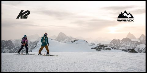 Dos personas suben una montaña nevada bajo un cielo nublado, con picos escarpados al fondo. Los logos de K2 y Wayback están en las esquinas superiores.