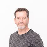 Un hombre con pelo corto y barba lleva una camisa de color gris oscuro con estampado y está de pie frente a un fondo blanco sencillo. Su expresión facial es tranquila y mira ligeramente hacia un lado.