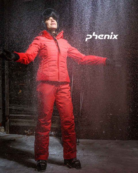 Una persona está de pie afuera en la nieve. Lleva un traje de esquí rojo brillante y guantes negros. Tiene los brazos extendidos y mira hacia arriba. Encima de ella se ve la palabra "Phenix". La escena sugiere que la persona está disfrutando del clima invernal.
