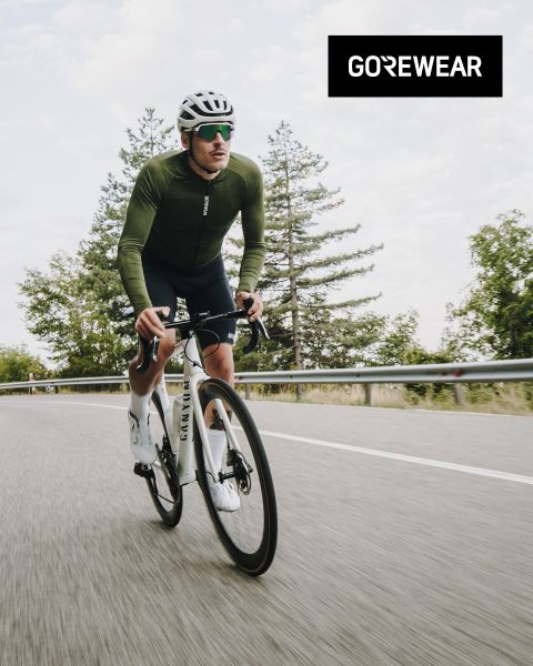 Ciclista con chaqueta verde y casco montando una bicicleta blanca en una carretera. En el fondo se ven árboles y un cielo nublado. El logo de "GOREWEAR" se ve en la esquina superior derecha.