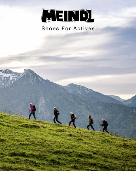 Un grupo de excursionistas con mochilas suben en fila una colina cubierta de hierba. En el fondo se ven montañas nevadas y un cielo nublado. En la parte superior de la imagen está el texto "MEINDL Shoes For Actives".