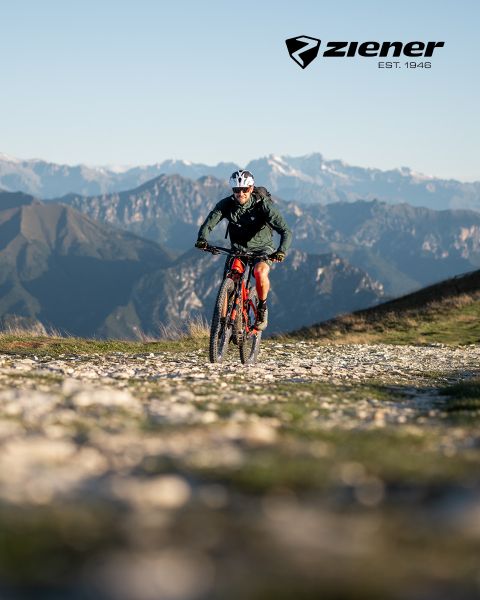 Un ciclista conduce su bicicleta de montaña roja por un camino de grava con el telón de fondo de montañas escarpadas. El cielo está despejado, el ciclista lleva casco y chaqueta verde. En la parte superior derecha está el logo de Ziener.