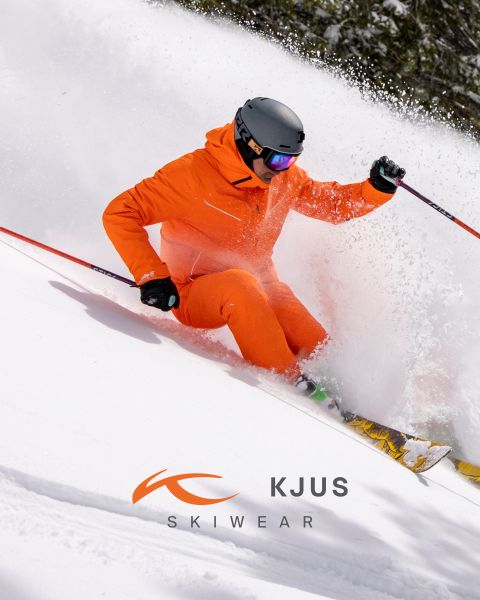 Un esquiador con un traje de esquí naranja brillante y un casco negro esquía a través de la nieve fresca, levantando nieve en polvo. El fondo está nevado y en la parte inferior de la imagen se ve el logotipo de KJUS Skiwear.