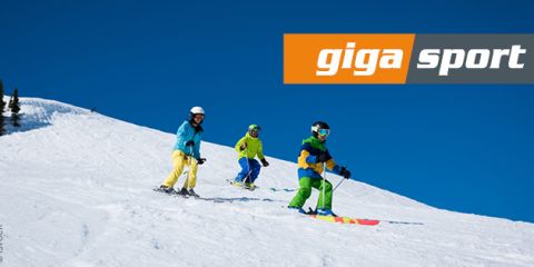 Drei Personen in farbenfroher Skiausrüstung fahren unter strahlend blauem Himmel einen verschneiten Hang hinunter. Oben rechts ist das Logo von „gigasport“ zu sehen.
