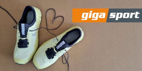 Ein Paar gelbe Sportschuhe mit schwarzen Details auf braunem Untergrund. Die Schnürsenkel bilden ein Herz. Das „Giga Sport“-Logo ist oben rechts zu sehen.