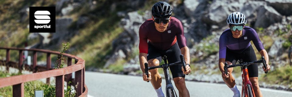 Dos ciclistas con equipamiento Sportful a juego suben por una carretera de montaña. Uno lleva un casco oscuro, el otro uno blanco. La carretera es sinuosa, con una ladera rocosa al fondo. En la esquina superior izquierda se ve el logotipo de "Sportful".