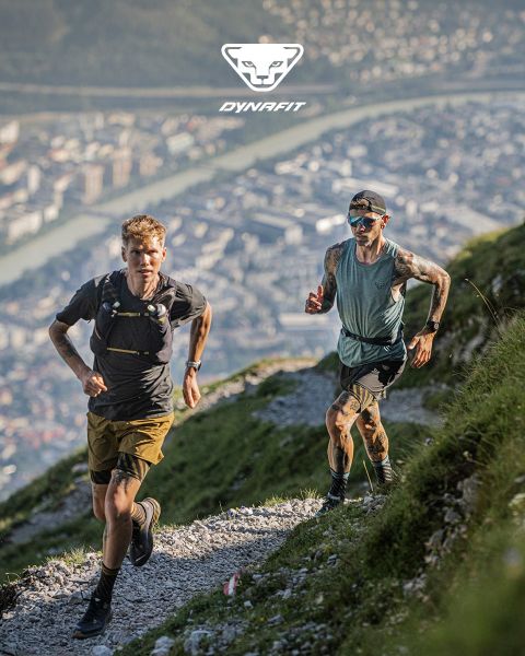 Dos personas corren por un sendero de montaña con una ciudad y un río al fondo. Uno lleva una camiseta negra y pantalones cortos, el otro una camiseta de tirantes azul y pantalones cortos. El logo de Dynafit está en la parte superior de la imagen.