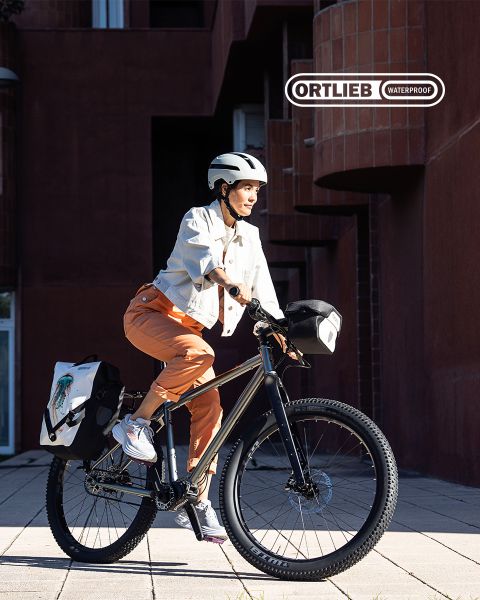 Una persona con casco y ropa casual anda en bicicleta con alforjas impermeables Ortlieb adjuntas, cerca de edificios modernos de ladrillo. El sol proyecta largas sombras y resalta su expresión facial concentrada.