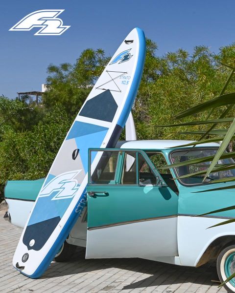 Un coche antiguo turquesa y blanco está estacionado con una tabla de paddle surf azul y blanca apoyada en él. En el fondo se ven árboles verdes y en la esquina superior izquierda hay un logo.