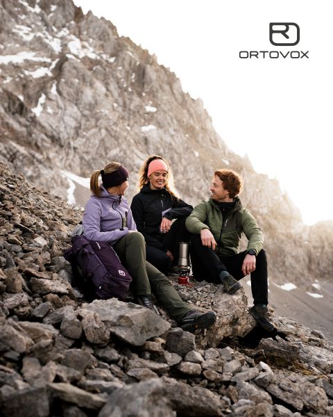 Tres personas con equipo para exteriores se sientan sonriendo y conversando en un terreno rocoso en una región montañosa. El sol se pone detrás de los picos. Una mochila y un termo están cerca. El logo de Ortovox está en la esquina superior derecha.