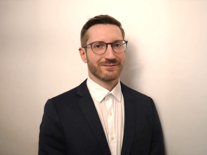 Una persona con gafas, camisa blanca y blazer oscuro está de pie frente a un fondo claro y sencillo.