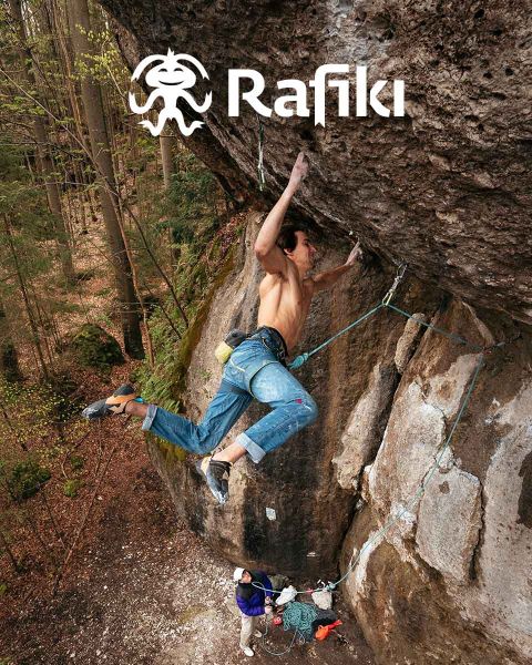 Un escalador en vaqueros y arnés de escalada asciende por una gran pared de roca con saliente, sujetándose a la roca con una mano. Debajo, una persona con una camisa blanca está de pie en el suelo en medio de su equipo de escalada. En la esquina superior está la palabra "Rafiki".