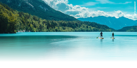 Dos personas practicando paddle surf en un tranquilo lago turquesa, rodeado de colinas boscosas y montañas a lo lejos bajo un cielo azul brillante con nubes dispersas.