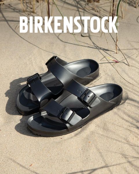 Una persona con sandalias Birkenstock beige está de pie junto a una fuente de agua en un exuberante jardín. El área está rodeada de vegetación y la palabra "BIRKENSTOCK" se ve claramente encima de la imagen.