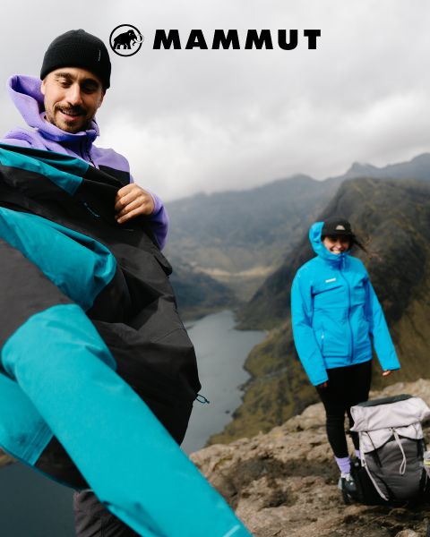 Dos excursionistas con equipo de exterior atraviesan un terreno rocoso bajo un cielo nublado, con una gran montaña al fondo. Ambos visten ropa de Mammut, y el logotipo de la marca se ve en la parte superior de la imagen.