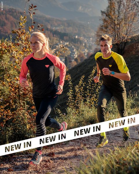 Dos personas en ropa deportiva corren por un sendero rocoso a través de un paisaje montañoso y boscoso. La luz del sol resalta el follaje otoñal. Pancartas blancas con la inscripción "NUEVO" en negritas letras negras atraviesan la parte inferior de la imagen.