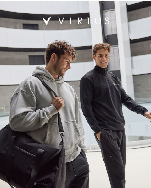 Dos hombres en ropa deportiva de Virtus, uno con una sudadera con capucha gris claro y una bolsa de lona negra, el otro con una chaqueta negra, sonríen y caminan juntos en un pasillo moderno. El logo de Virtus se puede ver encima de ellos.