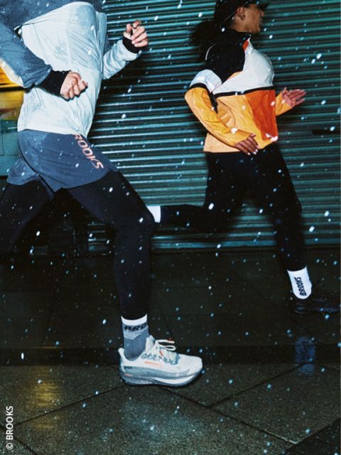Dos personas corriendo de noche en ropa deportiva, con gotas de lluvia visibles en el aire y aceras mojadas debajo de ellos. Ambos corredores llevan chaquetas y zapatillas de correr, y pasan rápidamente corriendo por una puerta de metal.