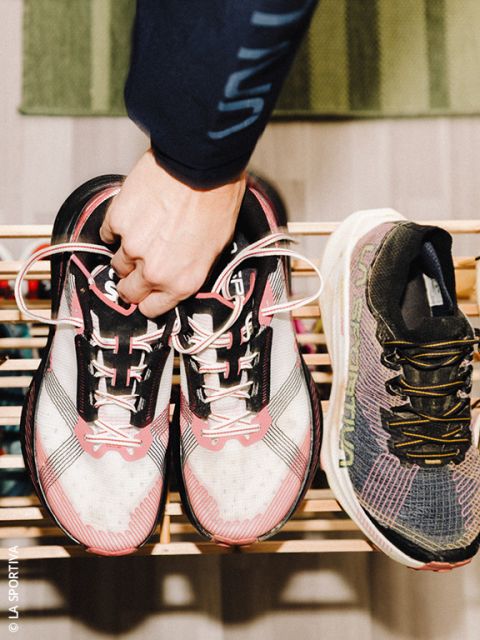 Una persona sostiene un par de zapatillas de correr de color blanco y rosa por los cordones sobre un zapatero de madera, junto a una sola zapatilla de correr de color negro y gris.