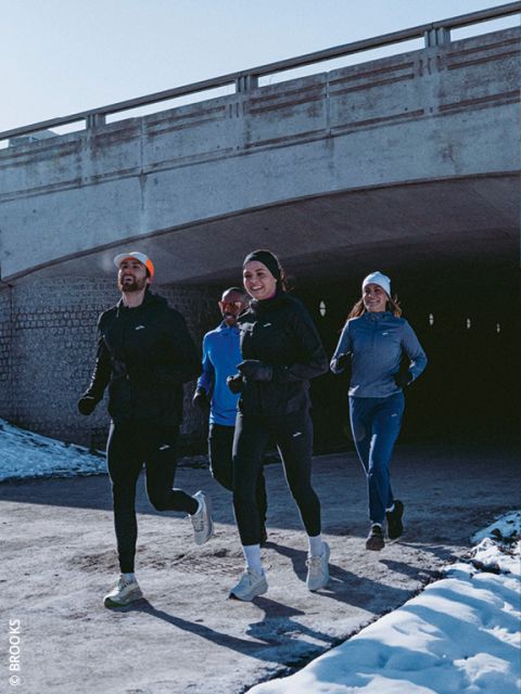 Cuatro personas con ropa de correr de invierno trotan juntas en un camino nevado bajo un puente de hormigón, sonriendo y disfrutando del ejercicio al aire libre.