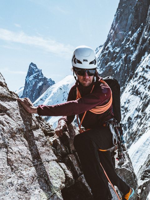 Un alpinista con casco, gafas de sol y equipo de escalada escala una ladera rocosa con picos nevados al fondo bajo un cielo despejado.