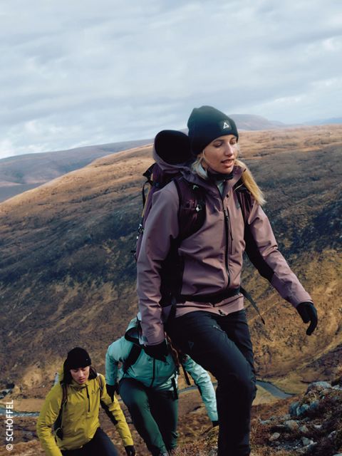 Tres excursionistas con equipo de exterior y mochilas escalan una ladera rocosa con colinas suaves y marrones al fondo bajo un cielo nublado. El excursionista de adelante lleva una chaqueta morada y un gorro.