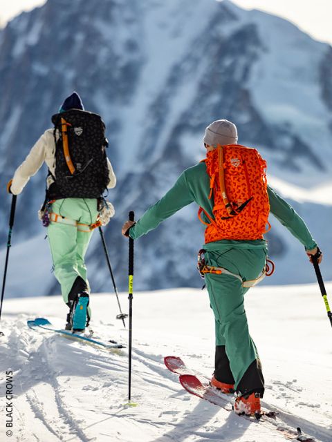 Dos personas con ropa de esquí colorida y mochilas suben una montaña nevada, apoyándose con bastones. En el fondo se ve un pico de montaña grande y escarpado.