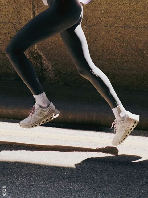 Una persona con zapatillas deportivas de color beige claro con suelas blancas y pantalones negros es fotografiada saltando con los pies en el suelo frente a un fondo oscuro.