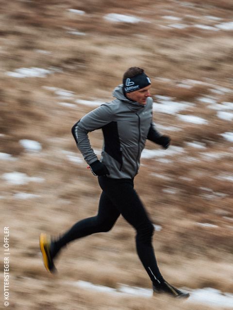 Una persona en ropa deportiva corre al aire libre por un camino cubierto de hierba borrosa con manchas de nieve, vestida con una chaqueta gris, leggings negros y una diadema.
