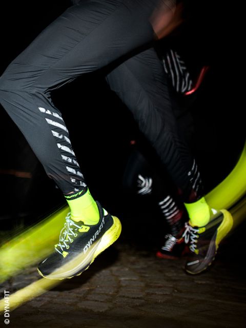 Un primer plano de dos personas corriendo de noche. El foco principal está en sus piernas, que visten pantalones deportivos negros, calcetines amarillos neón y zapatillas de correr DYNAFIT amarillas y negras, con el desenfoque de movimiento enfatizando la velocidad.