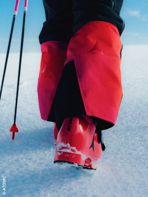 Una persona con pantalones de esquí de color marrón claro y botas de esquí grises está de pie en la nieve con bastones de esquí. Las botas de esquí tienen detalles rojos y un logo que dice "VAN DEER".