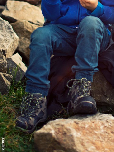 Dos chicos están sentados al aire libre sobre una roca. Un chico con una chaqueta azul sonríe mientras come un bocadillo, y el otro, con una chaqueta verde, mira hacia abajo y busca en su mochila. Ambos llevan botas de senderismo y mochilas.