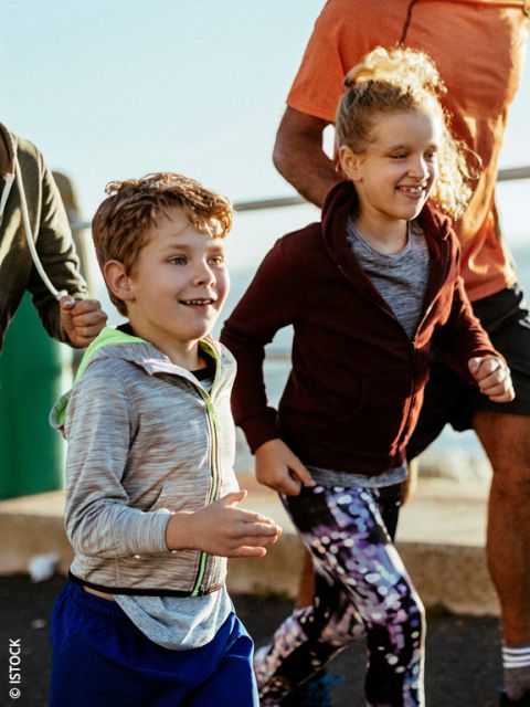 Dos niños sonrientes, un niño y una niña, trotan al aire libre con ropa deportiva. Adultos corren junto a ellos. La escena es soleada y enérgica, lo que sugiere una carrera familiar o grupal.
