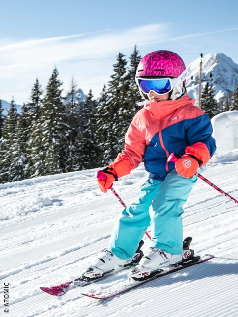 Un niño pequeño con un casco rosa brillante, gafas azules y un equipo de esquí colorido baja por una pista nevada, rodeado de pinos y montañas bajo un cielo azul despejado.