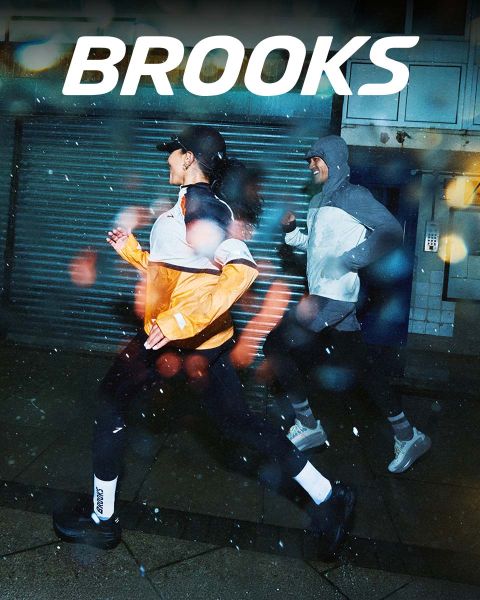 Un grupo de cinco personas corre enérgicamente por un paso de cebra. Llevan ropa deportiva colorida, incluyendo pantalones cortos y camisetas sin mangas. La escena es vibrante, con la palabra "BROOKS" estampada claramente encima de ellos.