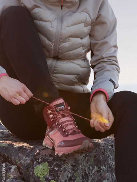 Una persona con una chaqueta acolchada clara y pantalones negros está sentada en una roca atándose los cordones de una bota de montaña marrón rojizo, mientras el sol de la tarde ilumina suavemente la escena.