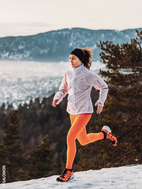 Una mujer con una chaqueta blanca y leggings de color naranja brillante corre por un sendero nevado con pinos y montañas al fondo. El cielo está nublado y el paisaje debajo de ella está parcialmente cubierto de nieve.