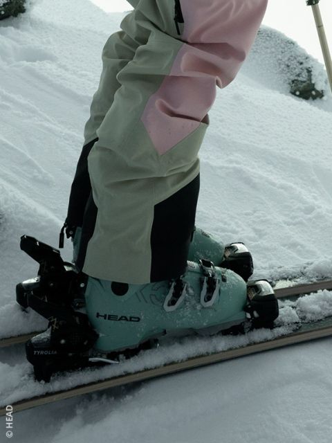 Una persona con botas de esquí HEAD verde menta y pantalones de esquí beige y rosa está de pie sobre esquíes en la nieve fresca.